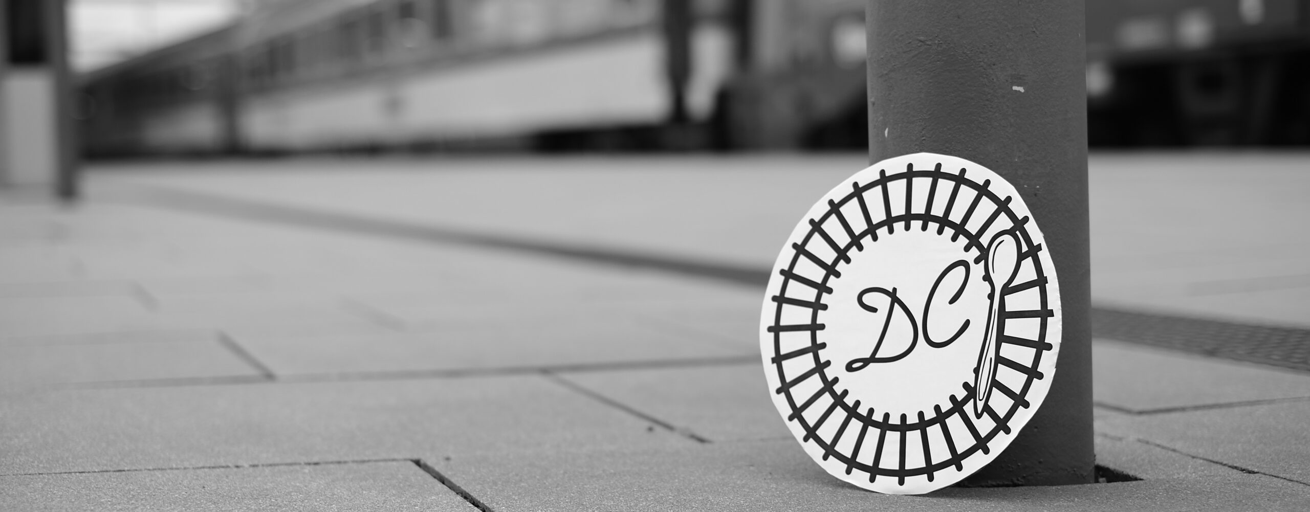 Black and white picture of the logo standing on a post on a railway station's platform. In the back a blurred train is visible.