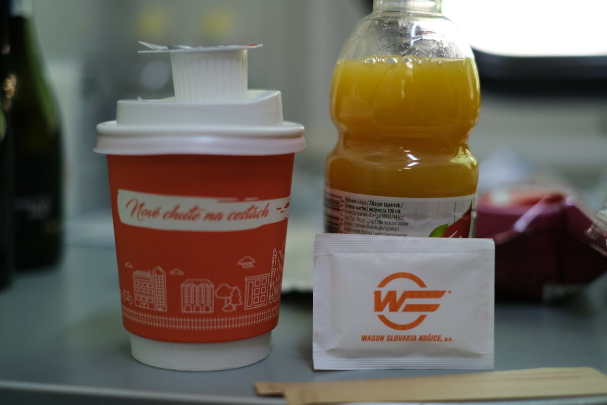 A paper coffee cup to go, a PET-bottle with orange juice and a package of sugar with the Wagon Slovakia logo on it. Brand colors are orange. Everthing stands oin a sleeper compartment's table.