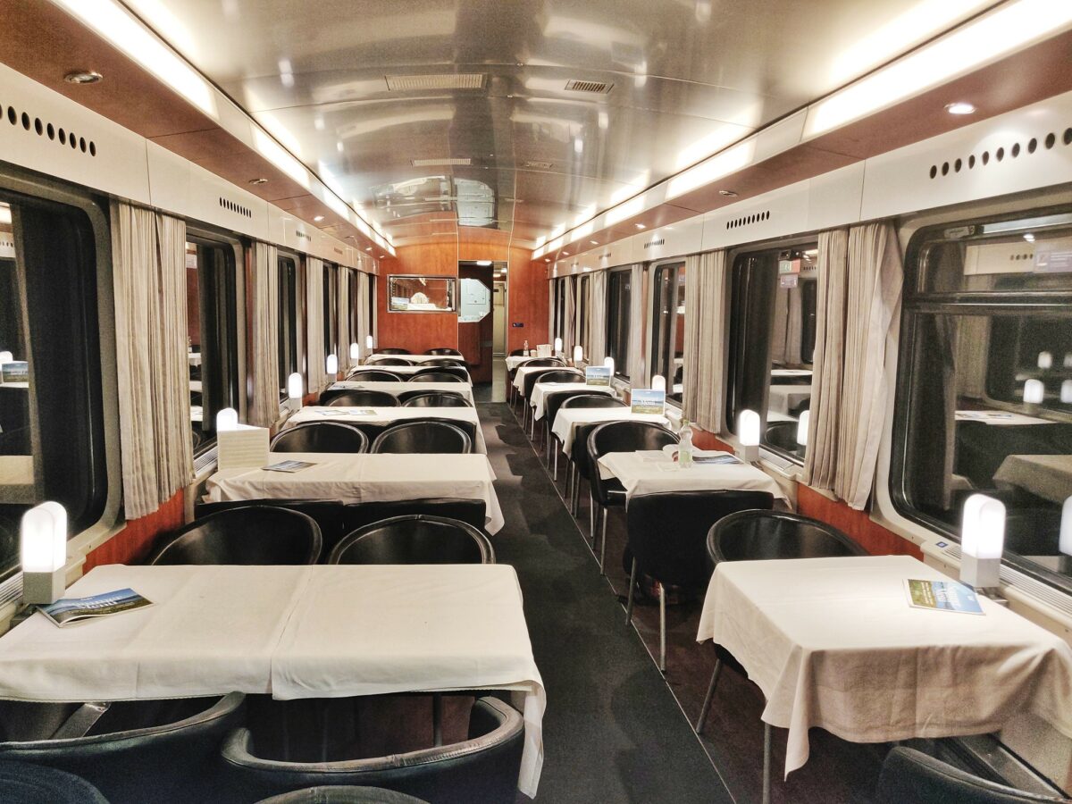 The inside of ÖBB's WRmz dining car. It's dark outside and lights are switched on. There are white table cloths and black leather chairs. No people are in the carriage.