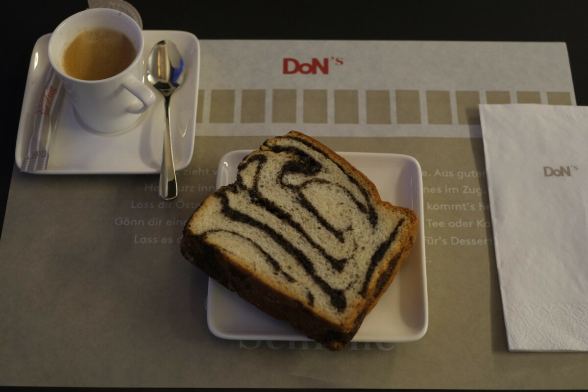Cup of espresso in white porcelain in the top left corner of the picture, centred a poppy seeds cake. The DoN's logo is visible on the branded table mat and tissue.