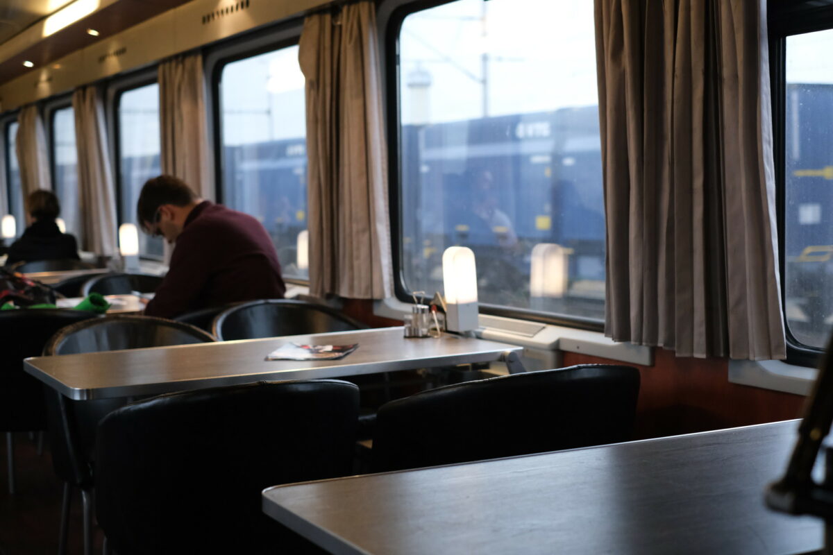 Picture of the inside of the dining car, with the table lamp switched on. Blurred in the Background there are people sitting on the balck leather chairs. No table cloths.