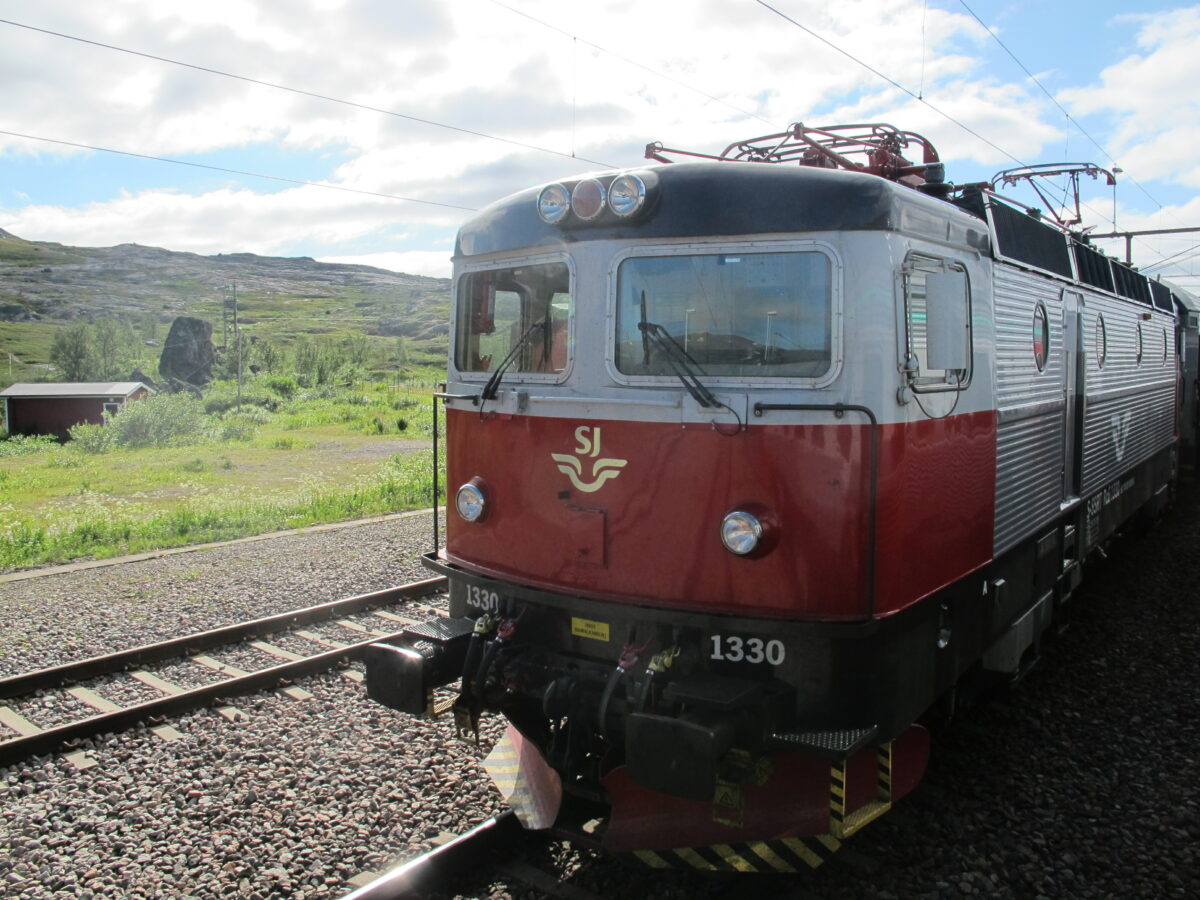 An SJ class 1330 in red and grey livery on tracks in nature. Soft hills and green grass are around.