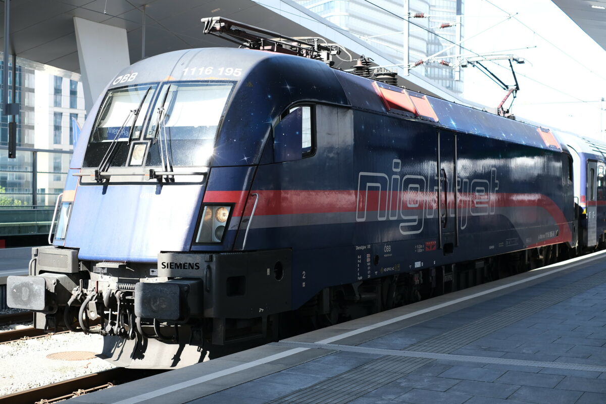 A ÖBB cass 1216 Taurus loco in a nightjet branded livery. It has a blue color with red and a grey stripe, also stars are visible on the blue background. The loco is waiting for departure on the platform of Vienna main train sation. The sun is shining brightly.